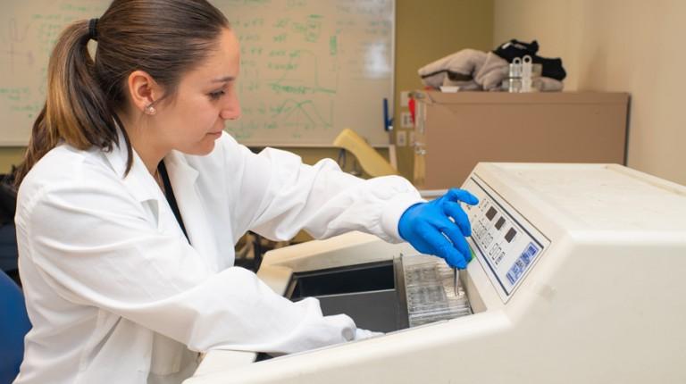 A student in a white lab coat works on a lab machine